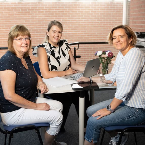 Drei Frauen sitzen gemeinsam an einem Tisch mit Laptop in einem Besprechungsraum.