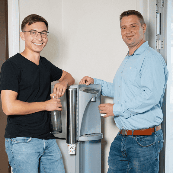 Zwei Mitarbeiter stehen an einem Wasserspender im Büro und füllen Trinkflaschen auf